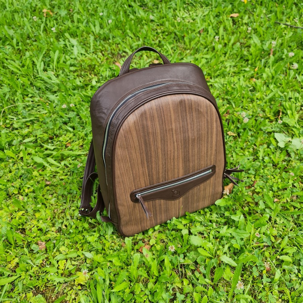 Nuo Backpack in brown leather with a wood-grain front panel and zipped front pocket, standing on grass.